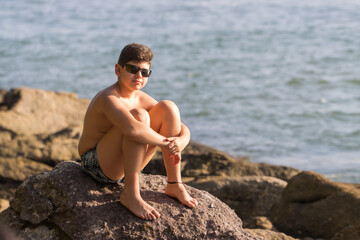 Fototapeta na wymiar Menino cheio de estilo sentado nas rochas da praia com o mar ao fundo e o sol iluminando parte de seu corpo e rosto que está usando óculos escuro. 