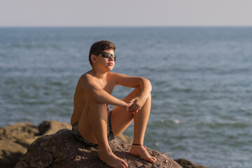 Fototapeta na wymiar Menino cheio de estilo sentado nas rochas da praia com o mar ao fundo e o sol iluminando parte de seu corpo e rosto que está usando óculos escuro. 