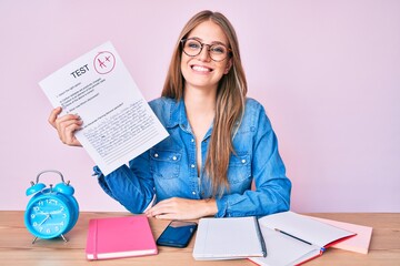 Young blonde girl showing passed exam sitting on the table looking positive and happy standing and smiling with a confident smile showing teeth