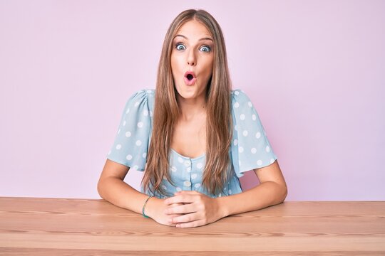 Young blonde girl wearing casual clothes sitting on the table scared and amazed with open mouth for surprise, disbelief face