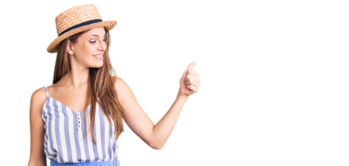Young beautiful blonde woman wearing summer hat looking proud, smiling doing thumbs up gesture to the side
