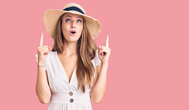 Young beautiful blonde woman wearing summer dress and hat amazed and surprised looking up and pointing with fingers and raised arms.