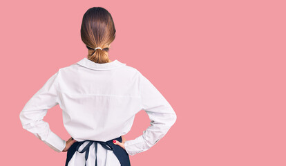 Beautiful brunette young woman wearing professional waitress apron standing backwards looking away...