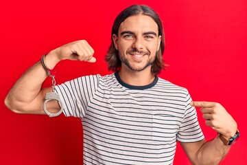 Young handsome man wearing prisoner handcuffs pointing finger to one self smiling happy and proud