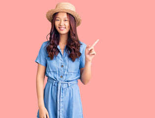 Young beautiful chinese girl wearing summer hat with a big smile on face, pointing with hand finger to the side looking at the camera.