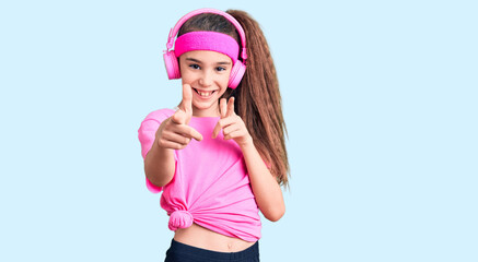 Obraz na płótnie Canvas Cute hispanic child girl wearing gym clothes and using headphones pointing fingers to camera with happy and funny face. good energy and vibes.