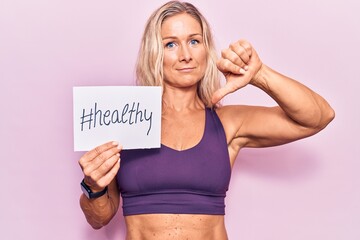 Middle age caucasian blonde woman wearing sportswear holding healthy banner with angry face, negative sign showing dislike with thumbs down, rejection concept