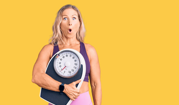 Middle age fit blonde woman wearing sports clothes holding weighing machine scared and amazed with open mouth for surprise, disbelief face
