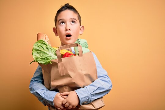 Adorable toddler holding paper bag with food standing over isolated yellow background