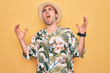 Young handsome man with blue eyes on vacation wearing summer florar shirt and hat crazy and mad shouting and yelling with aggressive expression and arms raised. Frustration concept.