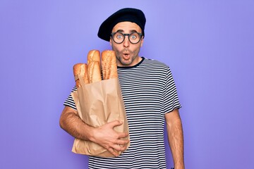 Young handsome baker man with blue eyes wearing french beret holding bag with bread scared in shock...