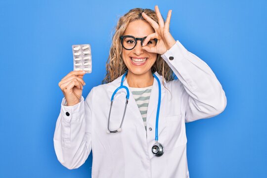 Young beautiful blonde cardiologist woman wearing stethoscope holding plastic heart smiling happy doing ok sign with hand on eye looking through fingers