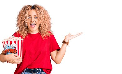 Young blonde woman with curly hair eating popcorn celebrating victory with happy smile and winner expression with raised hands