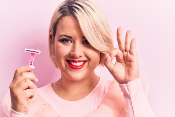 Young beautiful blonde plus size woman holding depilation razor over isolated pink background doing ok sign with fingers, smiling friendly gesturing excellent symbol