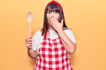 Young plus size woman wearing apron holding wooden spoon covering mouth with hand, shocked and afraid for mistake. surprised expression