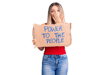 Young beautiful blonde woman holding power to the people banner covering mouth with hand, shocked...