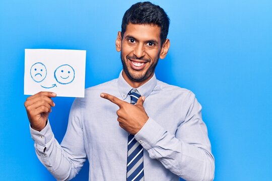 Young latin man holding sad to happy emotion paper smiling happy pointing with hand and finger