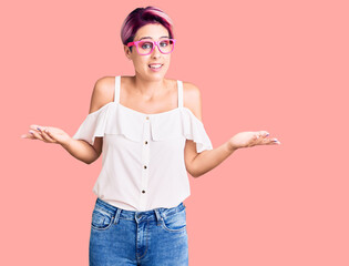 Young beautiful woman with pink hair wearing casual clothes and glasses clueless and confused expression with arms and hands raised. doubt concept.
