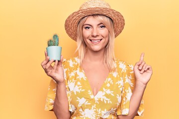 Beautiful blonde woman wearing summer hat holding cactus plant pot over yellow background smiling happy pointing with hand and finger to the side