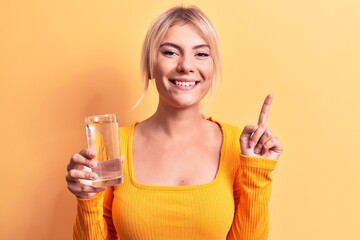 Young beautiful blonde woman drinking glass of water over isolated yellow background smiling with an idea or question pointing finger with happy face, number one