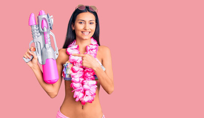 Young beautiful latin girl wearing bikini and hawaiian lei holding water gun smiling happy pointing...