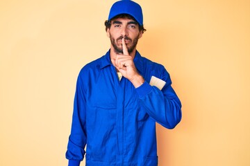 Handsome young man with curly hair and bear wearing builder jumpsuit uniform asking to be quiet with finger on lips. silence and secret concept.