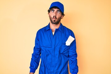 Handsome young man with curly hair and bear wearing builder jumpsuit uniform making fish face with lips, crazy and comical gesture. funny expression.