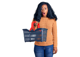 Beautiful african american woman holding supermarket shopping basket scared and amazed with open mouth for surprise, disbelief face