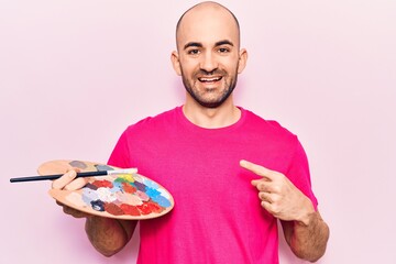 Young handsome bald man painting using paintbrush and palette smiling happy pointing with hand and finger