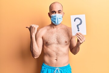 Fototapeta premium Young handsome bald man wearing swimwear and medical mask holding question mark pointing thumb up to the side smiling happy with open mouth