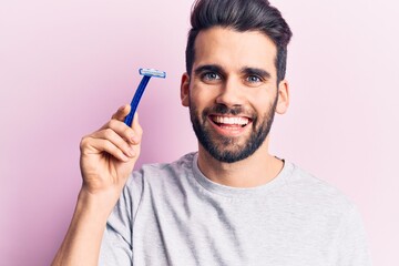 Obraz premium Young handsome man with beard holding razor looking positive and happy standing and smiling with a confident smile showing teeth