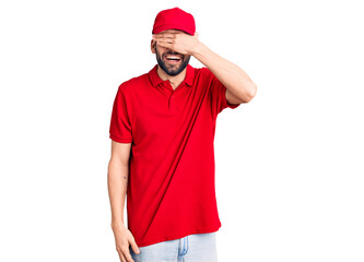 Young handsome man with beard wearing delivery uniform smiling and laughing with hand on face covering eyes for surprise. blind concept.