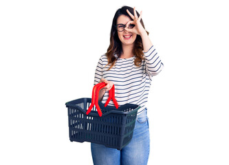 Beautiful young brunette woman holding supermarket shopping basket smiling happy doing ok sign with hand on eye looking through fingers