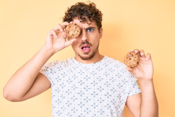 Young caucasian man with curly hair holding cookie in shock face, looking skeptical and sarcastic, surprised with open mouth