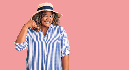 Young african american plus size woman wearing summer hat smiling doing phone gesture with hand and fingers like talking on the telephone. communicating concepts.