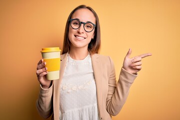 Young beautiful businesswoman drinking cup of takeaway coffee over yellow background very happy pointing with hand and finger to the side