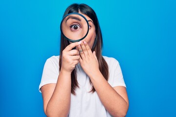 Young beautiful brunette woman using magnifying glass over isolated blue background covering mouth with hand, shocked and afraid for mistake. Surprised expression