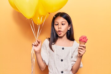 Adorable hispanic child girl holding balloons eating ice cream standing over isolated yellow...