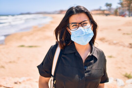 Young beautiful brunette woman wearing medical mask and glasses walking at the beach