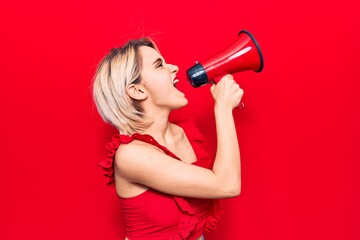 Young beautiful blonde woman with angry expression. Screaming loud using megaphone standing over isolated red background