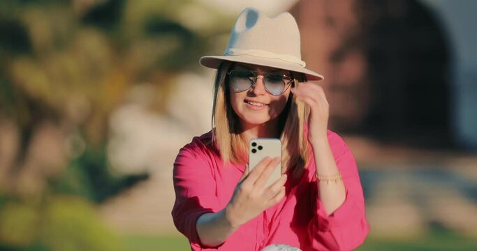 4K Smiling attractive blogger woman in stylish casual outfit making a selfie or video call to boyfriend or parents in a park at beautiful golden sunset light. Concept of technology wireless connection