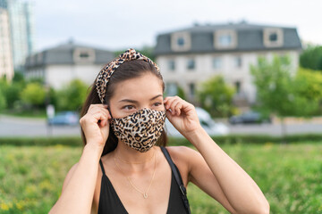 Asian woman putting on COVID-19 face mask wearing leopard fabric pattern masks matching fashion outfit outside in city for coronavirus. Summer lifestyle outdoor. Corona virus.
