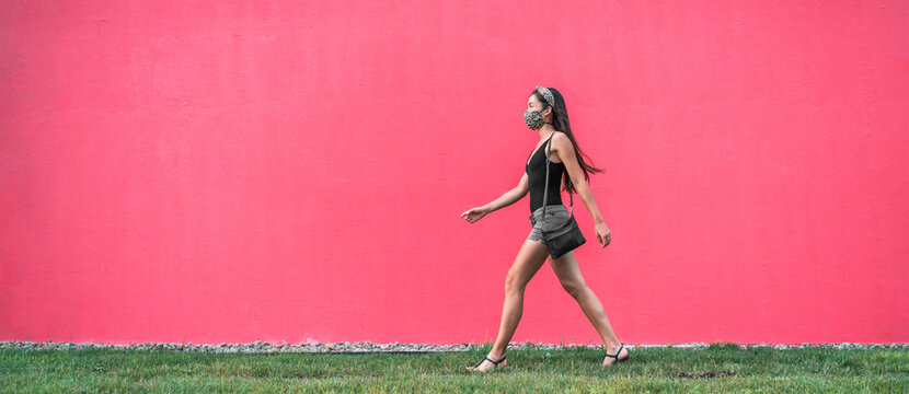 COVID-19 mask wear in city girl walking wearing mask as coronavirus prevention outside in city against street pink wall banner background. Corona virus new lifestyle.