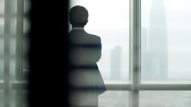 rear view of a mature asian business man standing by the window in office looking out at modern buildings 