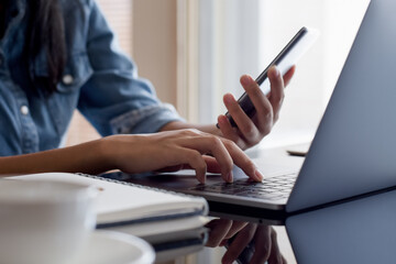 Young asian business woman use mobile smart phone and work on laptop computer with notebook on the desk. Student online education, online working concept.