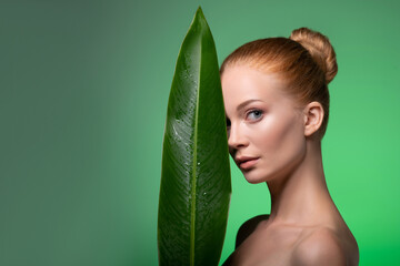 Portrait of healthy woman with half face is hidden behind a big tropical palm leaf on green...