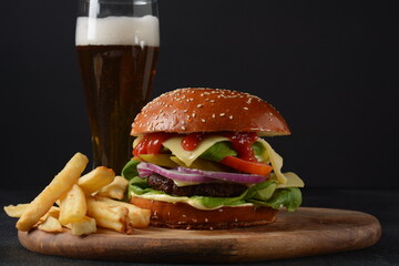 Homemade burger with grilled beef, cheese, onion, tomato, and green salad, sauce, with pickles and French fries,and a glass of beer in the background.
