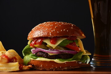 Homemade burger with grilled beef, cheese, onion, tomato, and green salad, sauce, with pickles and French fries,and a glass of beer in the background.
