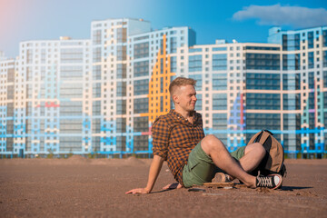 Fototapeta na wymiar A young man in a checked shirt and carrying a backpack sits on the sand with a view of multi-storey new buildings in multicolored colors, housing for young families