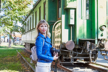 Attractive beautiful young girl who is female traveller in a trip standing near near the railway car and holding modern mirror camera in autumn park